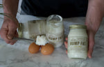 Person using a spoon to scoop cream from a jar labeled 'Hump Fat' on a marble surface with eggs.