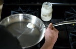 Person spooning a white substance into a frying pan on a stove with a jar of the same substance in the background.